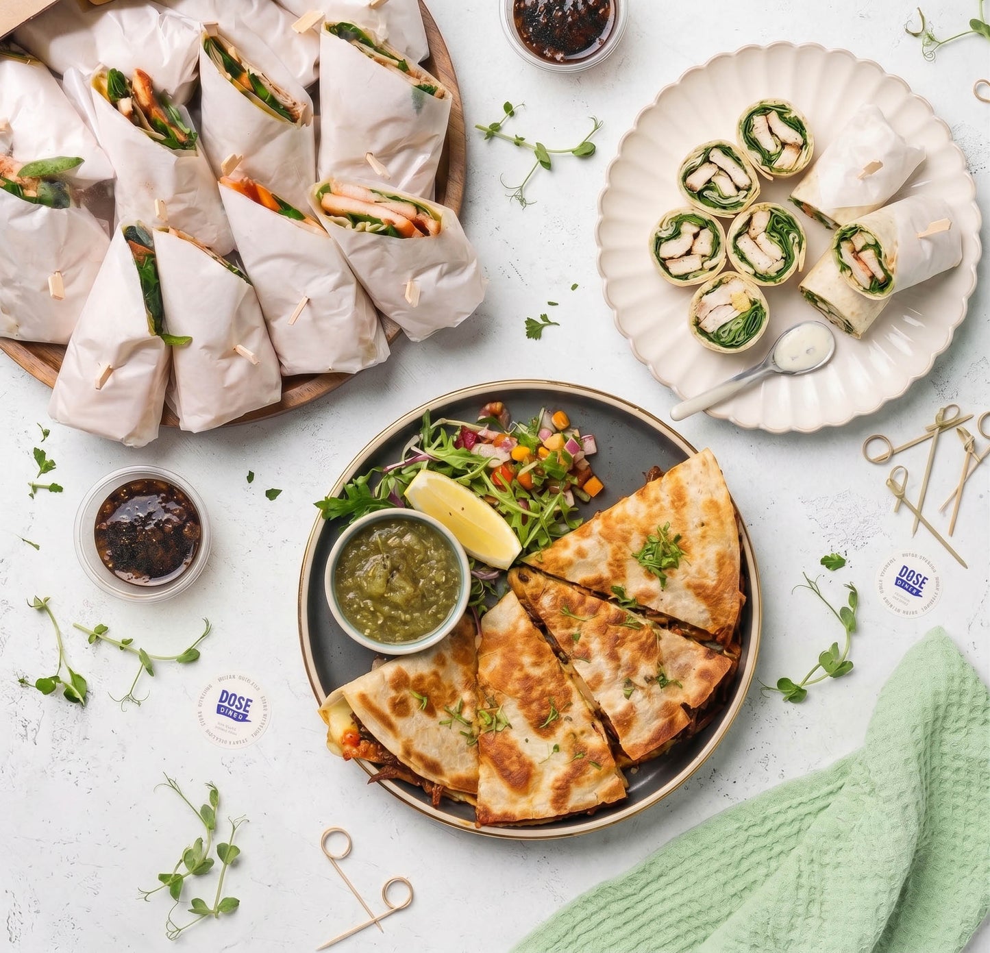 A styled overhead food spread on a white surface, featuring wrapped wraps on a wooden board, rolled green salad wraps on a scalloped plate, and a large plate of stuffed flatbread wedges with dips and garnish. Small branded stickers reading "Breeze" are scattered around.