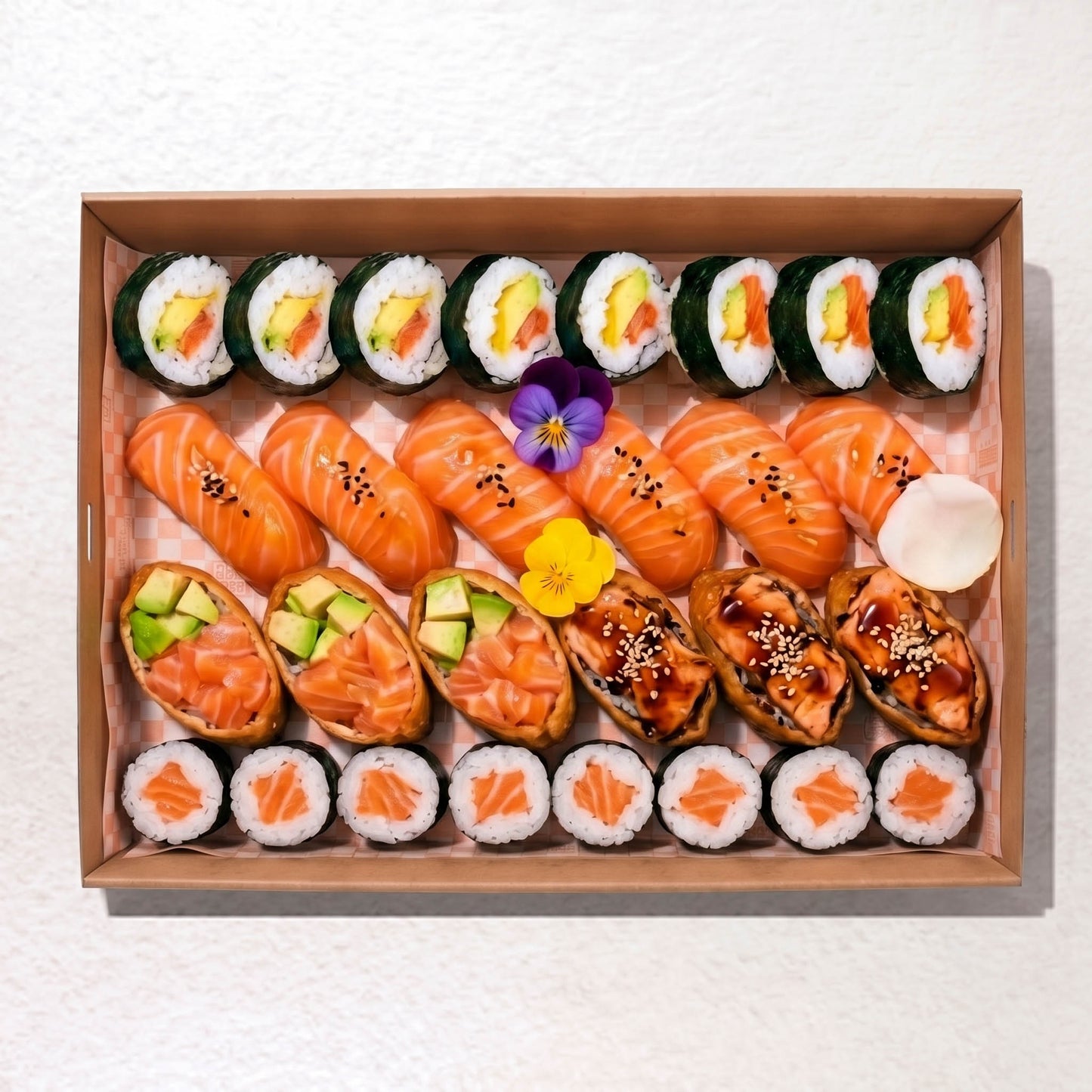 A top-down view of a sushi box containing salmon maki, avocado salmon nigiri, tuna maki, and other assorted sushi rolls, arranged neatly in a brown cardboard tray on a white textured background.