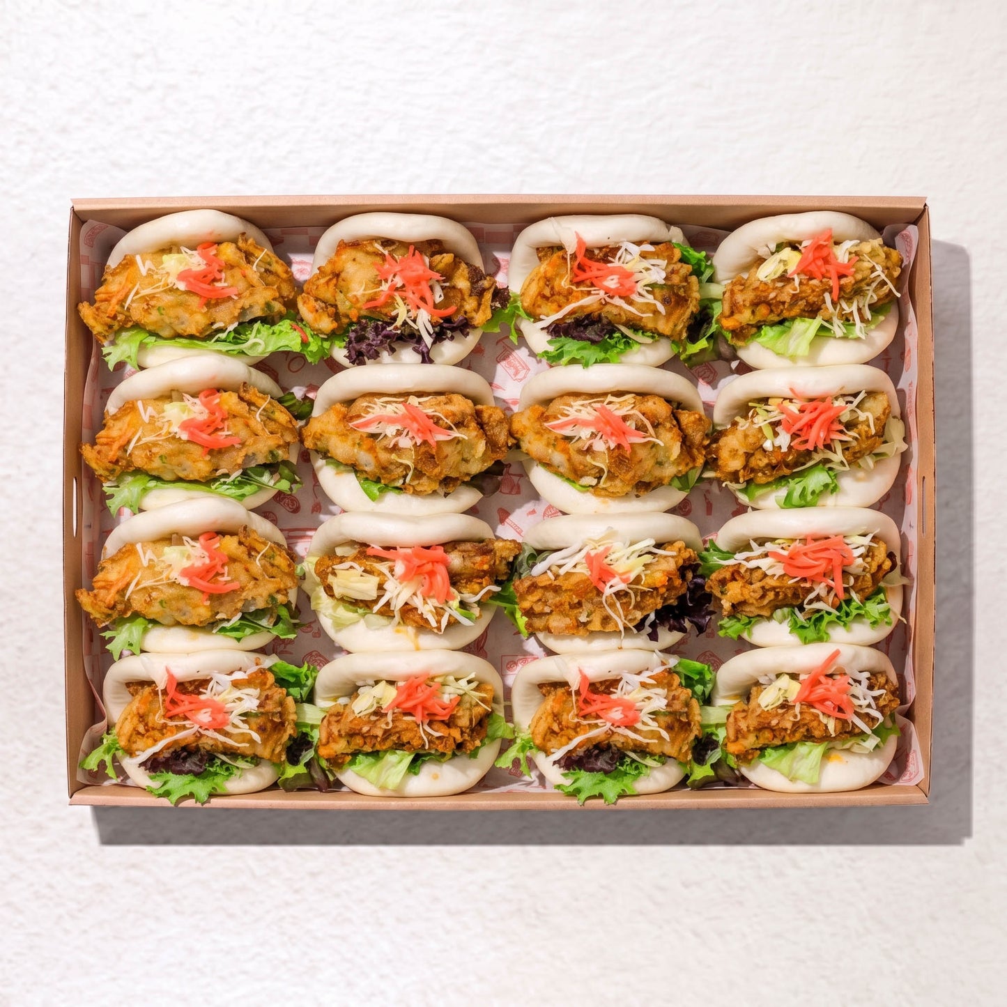 A top-down view of a rectangular cardboard tray holding 15 mini sandwich wraps filled with fried chicken, lettuce, shredded cabbage, and pickled ginger on a white textured background. from Double Dribble Catering, Christchurch