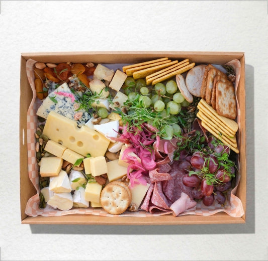 A top-down view of a gourmet charcuterie board arranged neatly inside a square cardboard tray, featuring various cheeses, crackers, grapes, salami, and greens on a light textured background.