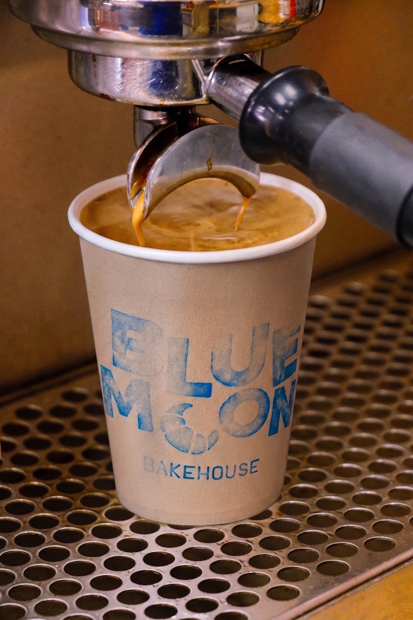 A close-up of espresso being poured into a paper cup printed with the words 'BLUE MEON BAKEHOUSE' in blue lettering. The cup sits on a metal drip tray beneath an espresso machine portafilter.