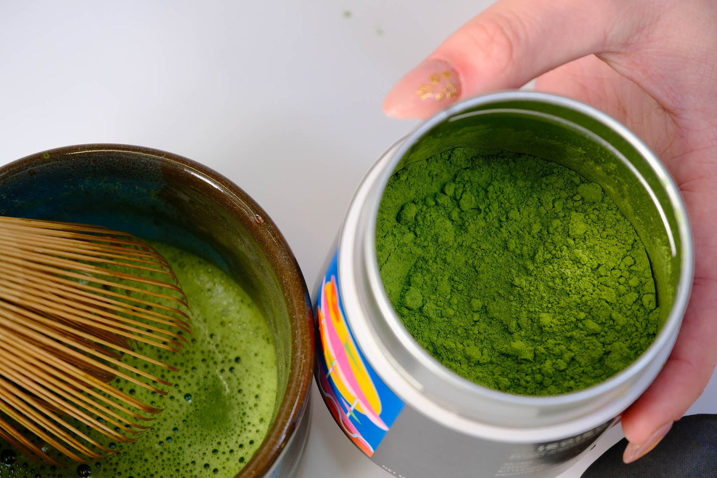 Open tin of vivid green Komorebi matcha beside a whisked bowl