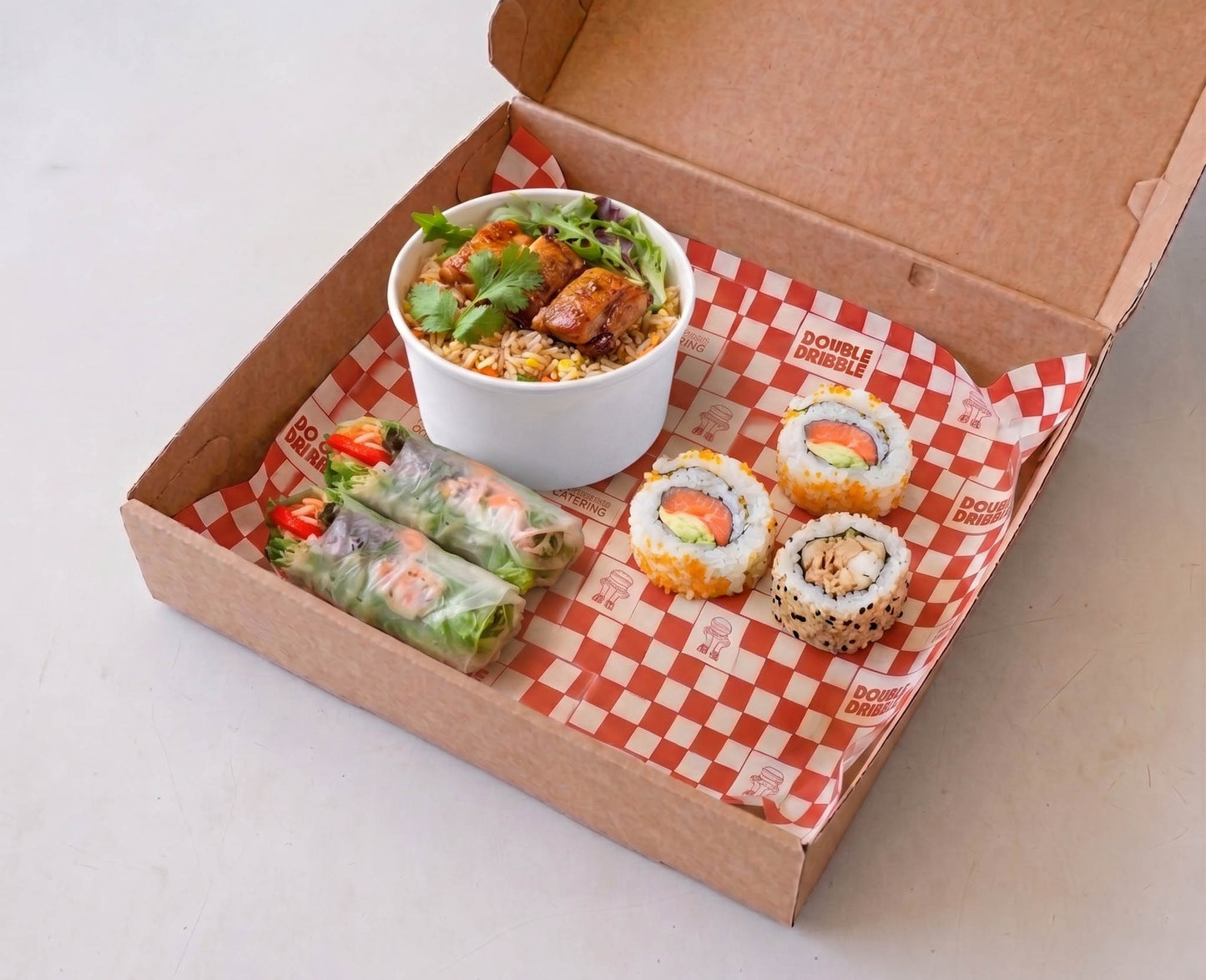 A cardboard take-out box lined with red-and-white checkered paper, containing a white cup of salad with grilled pieces, two spring rolls, and four sushi rolls, photographed on a light neutral surface.