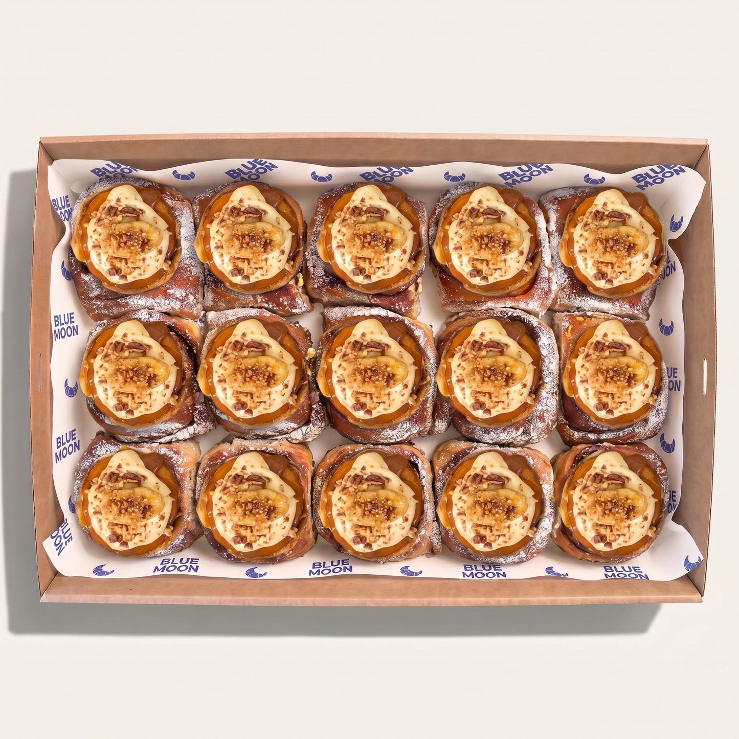 A top-down view of a cardboard bakery box filled with 12 baked pastries, each topped with a golden-brown caramelized custard or cream and a sprinkle of powdered sugar. The box is lined with white paper printed with the brand name 'Blue Moon'.