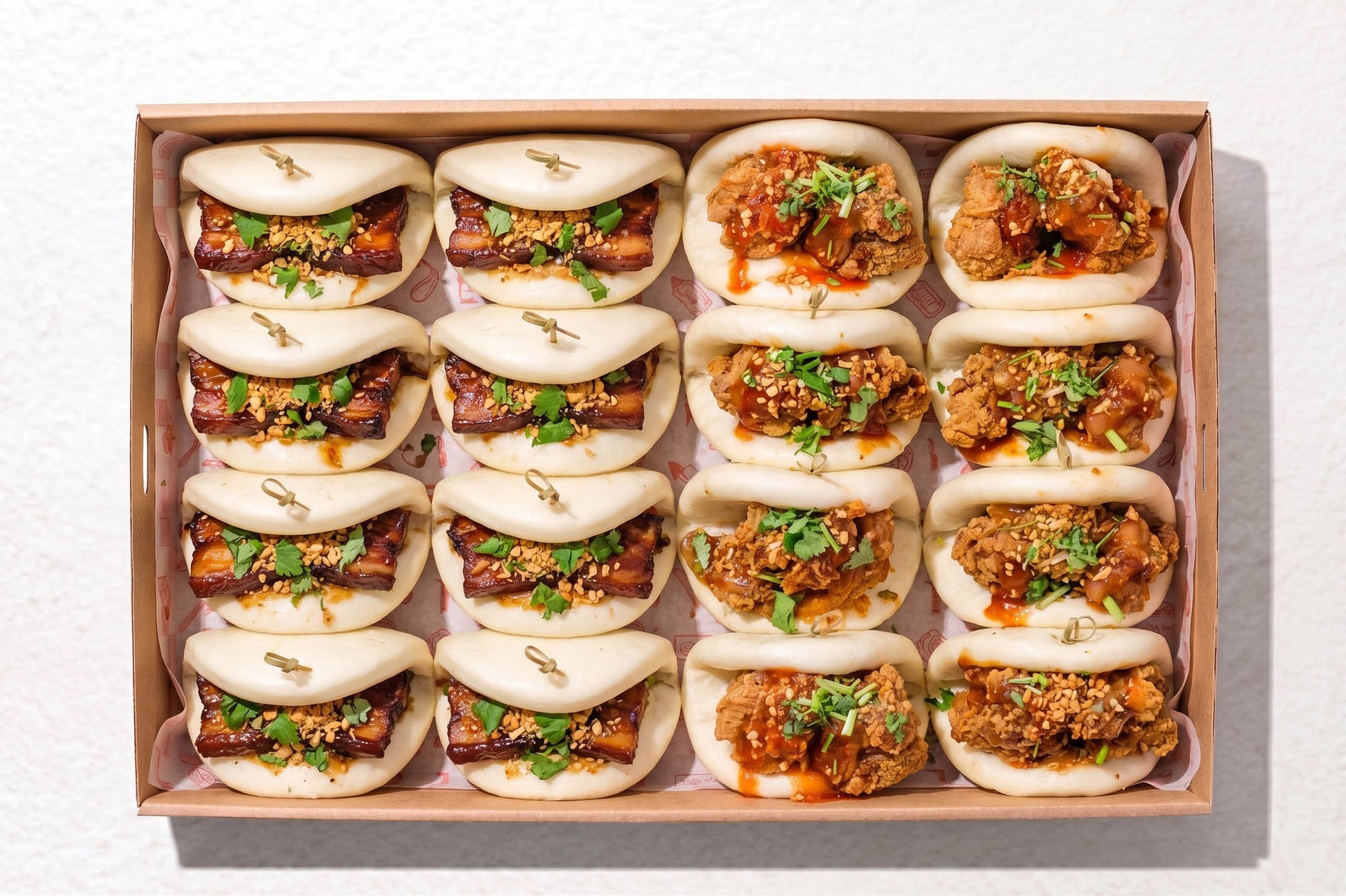 A top-down view of a wooden box containing 15 mini bao buns filled with various savory meats, sesame seeds, and green scallions, set against a clean white background.