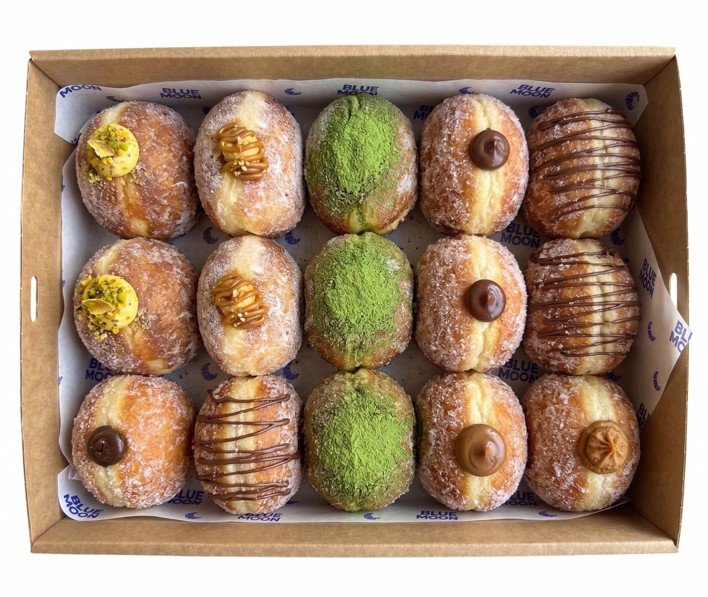 A top-down view of a cardboard box containing 12 assorted donuts. The donuts include sugar-dusted varieties topped with pistachio, walnuts, chocolate drizzle, and matcha green powder, as well as glazed donuts with chocolate toppings. Blue Moon Bakehouse, 70 Riccarton rd, christchurch, Double Dribble Catering