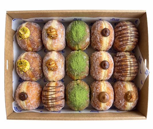 A top-down view of a cardboard box containing 12 assorted donuts. The donuts include sugar-dusted varieties topped with pistachio, walnuts, chocolate drizzle, and matcha green powder, as well as glazed donuts with chocolate toppings. Blue Moon Bakehouse, 70 Riccarton rd, christchurch, Double Dribble Catering