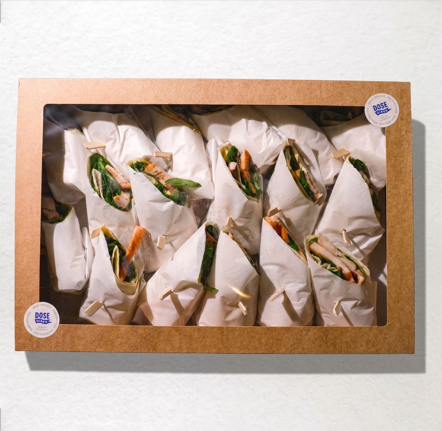 A square cardboard window box containing multiple wrapped food items, likely wraps or burritos, arranged inside on a white background. Two round stickers on the box read 'DOSE FOOD'.