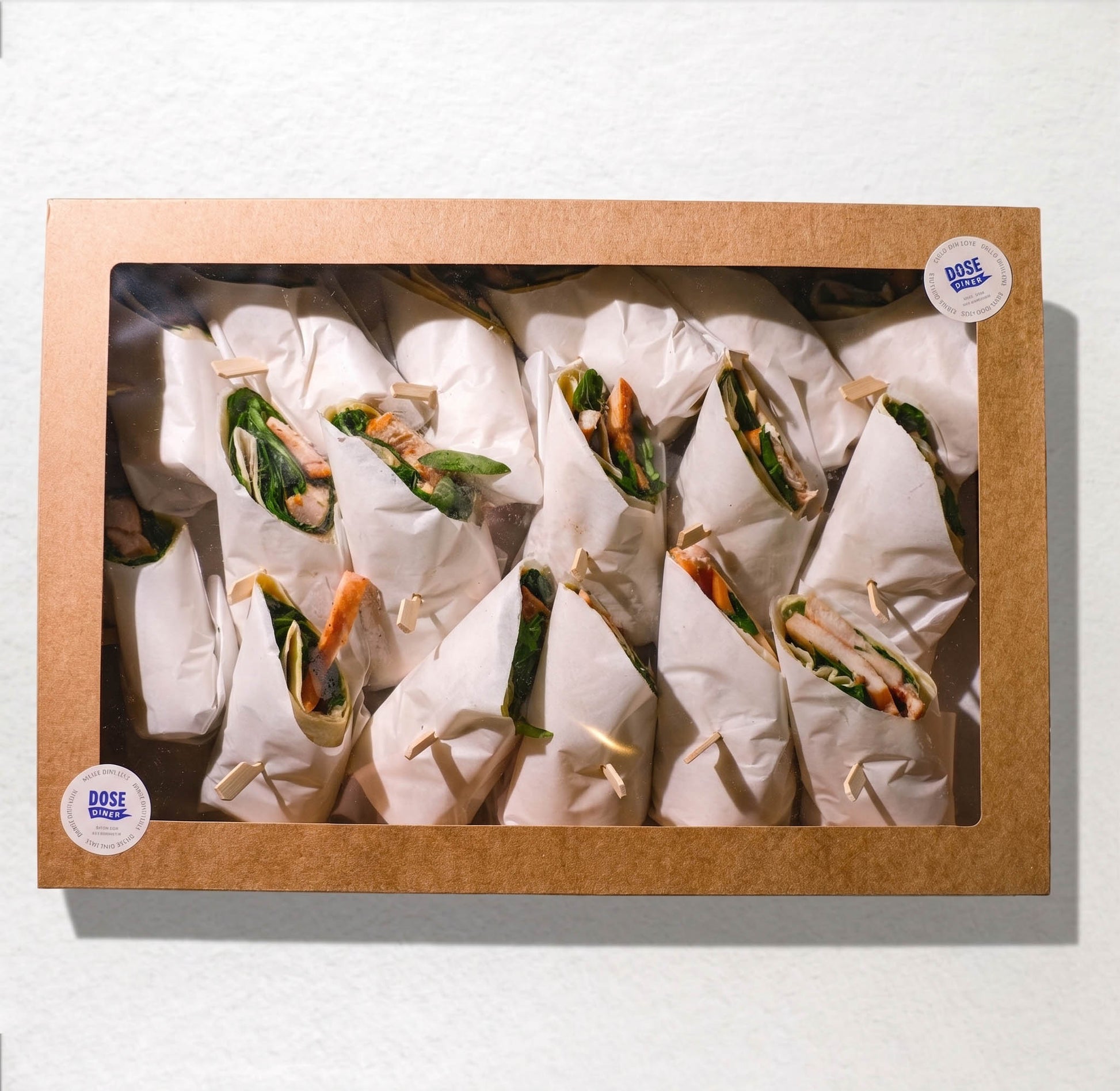 A square cardboard window box containing multiple wrapped food items, likely wraps or burritos, arranged inside on a white background. Two round stickers on the box read 'DOSE FOOD'.