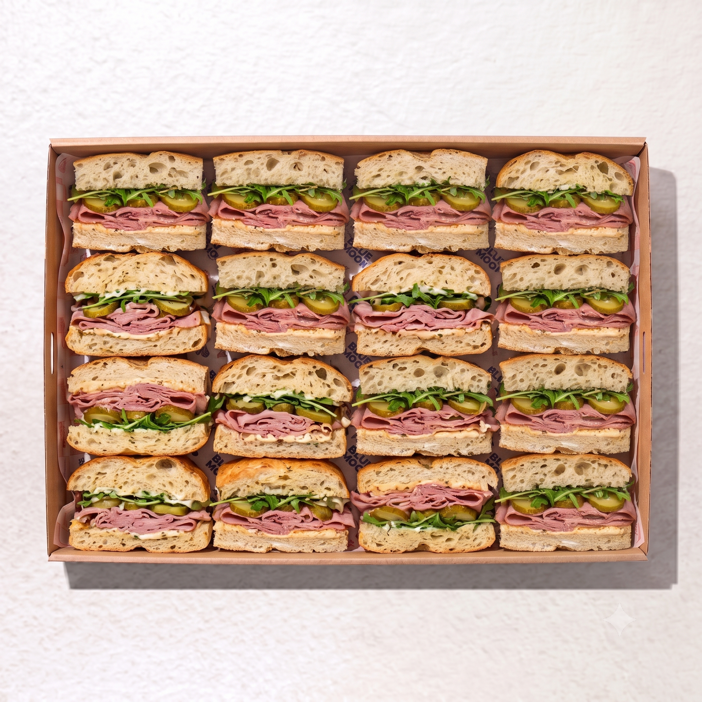 A top-down view of a cardboard tray holding 18 sliced open-faced sandwiches arranged in a 3x6 grid on a white textured surface.