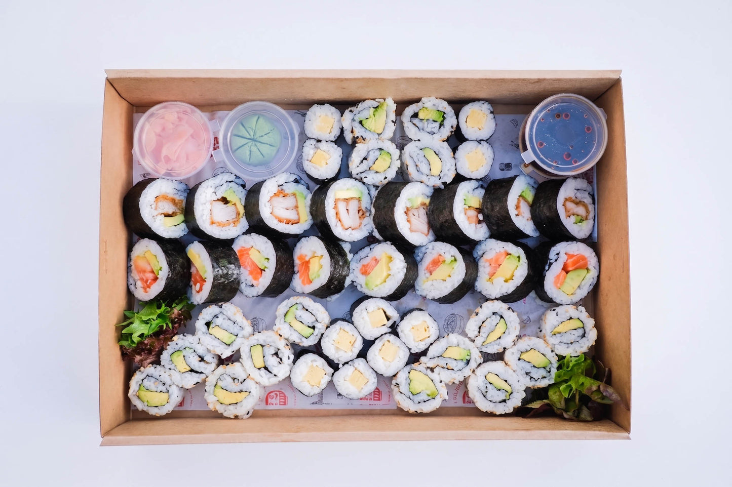A wooden box filled with assorted sushi rolls and small containers of dipping sauces, photographed from above on a plain white background.