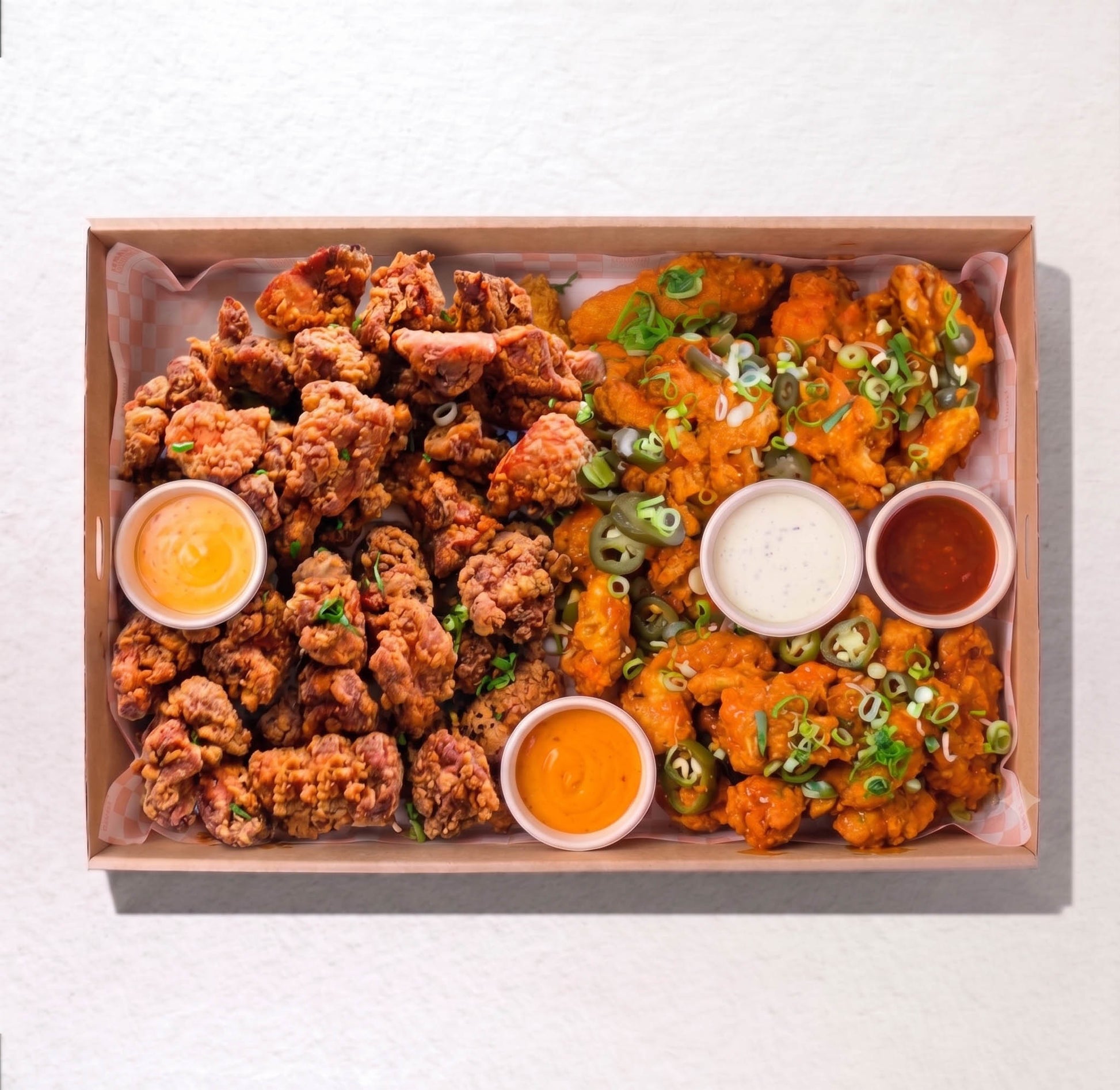 A top-down view of a cardboard food tray filled with two styles of fried chicken: crispy golden-brown fried pieces on the left and orange-battered pieces on the right with green onions, accompanied by four dipping sauces. by Boom Boom Chicken, in the Little High eatery, Christchurch 