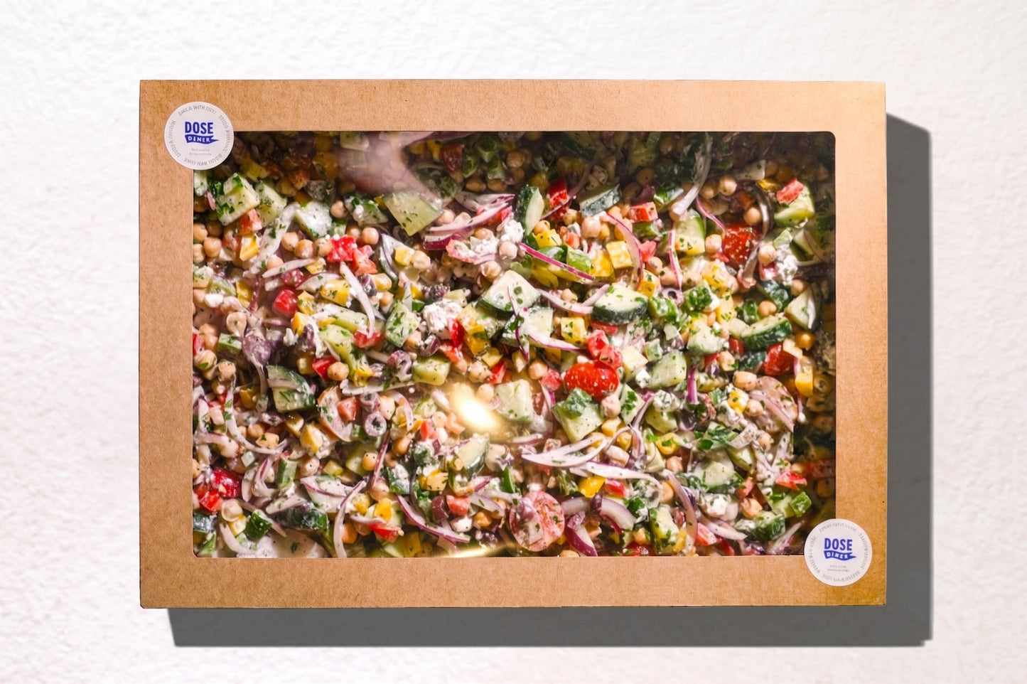 A square framed art print featuring a colorful fresh salad, mounted on a light beige textured wall. The print is housed in a natural brown paper box frame with two circular white Dose Diner logo, catering made in Christchurch