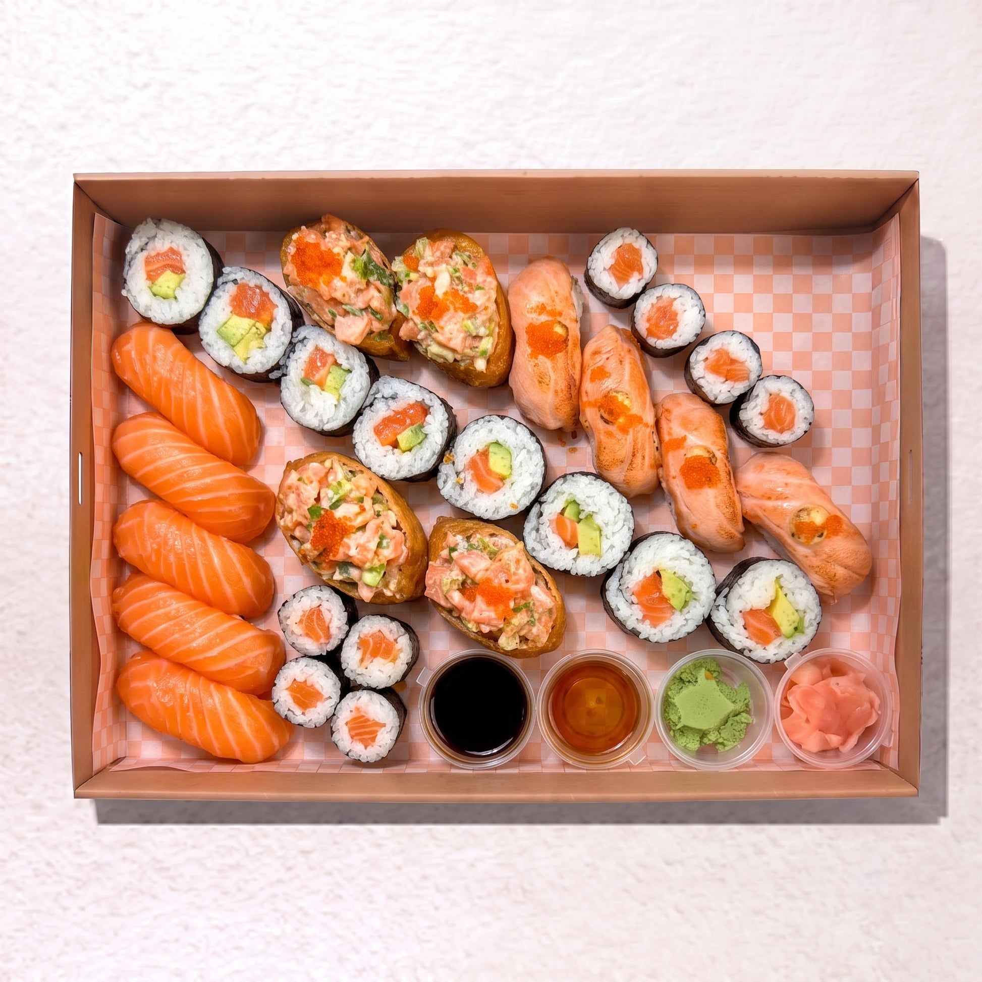 A sushi platter arranged in a rectangular cardboard tray on a light textured surface. The set includes salmon nigiri, maki rolls, salmon and avocado stuffed sushi pieces, and small dipping sauces in soy sauce, teriyaki sauce, and wasabi, with pickled ginger on the side.