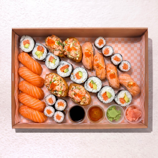 A sushi platter arranged in a rectangular cardboard tray on a light textured surface. The set includes salmon nigiri, maki rolls, salmon and avocado stuffed sushi pieces, and small dipping sauces in soy sauce, teriyaki sauce, and wasabi, with pickled ginger on the side.